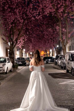 A bride in an elegant white dress walks beneath glowing Bonn cherry blossom trees on a quiet street in Bonn, Germany, captured as a romantic wedding moment by Vidora Studio.