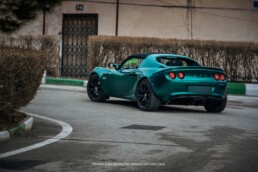 A metallic emerald green Lotus Elise with a black soft top is parked on a curved road, with a building and dry bushes in the background.