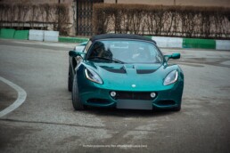 A metallic green Lotus Elise with its soft top on, photographed from the front angle as it turns on a damp, curved road.