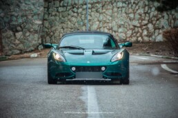 A head-on shot of a metallic green Lotus Elise on a wet road, with its headlights illuminated against a rustic stone wall background.