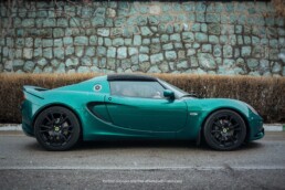 A metallic green Lotus Elise with black wheels and a soft top, photographed from the side against a rustic stone wall and dry hedges.