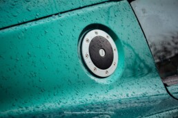 A detailed shot of the motorsports-style fuel cap on a metallic green car. The car's paint is covered in shimmering water droplets from rain.