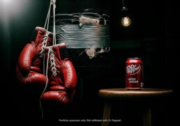 In a dark gym setting, a can of Dr Pepper Zero Sugar sits on a wooden stool, illuminated by a single lightbulb, with red boxing gloves hanging next to a worn punching bag.