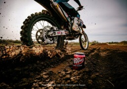 From a low angle, a can of Dr Pepper Zero Sugar sits in the mud of a dirt track while the rear wheel of a motocross bike sprays mud and debris past it.
